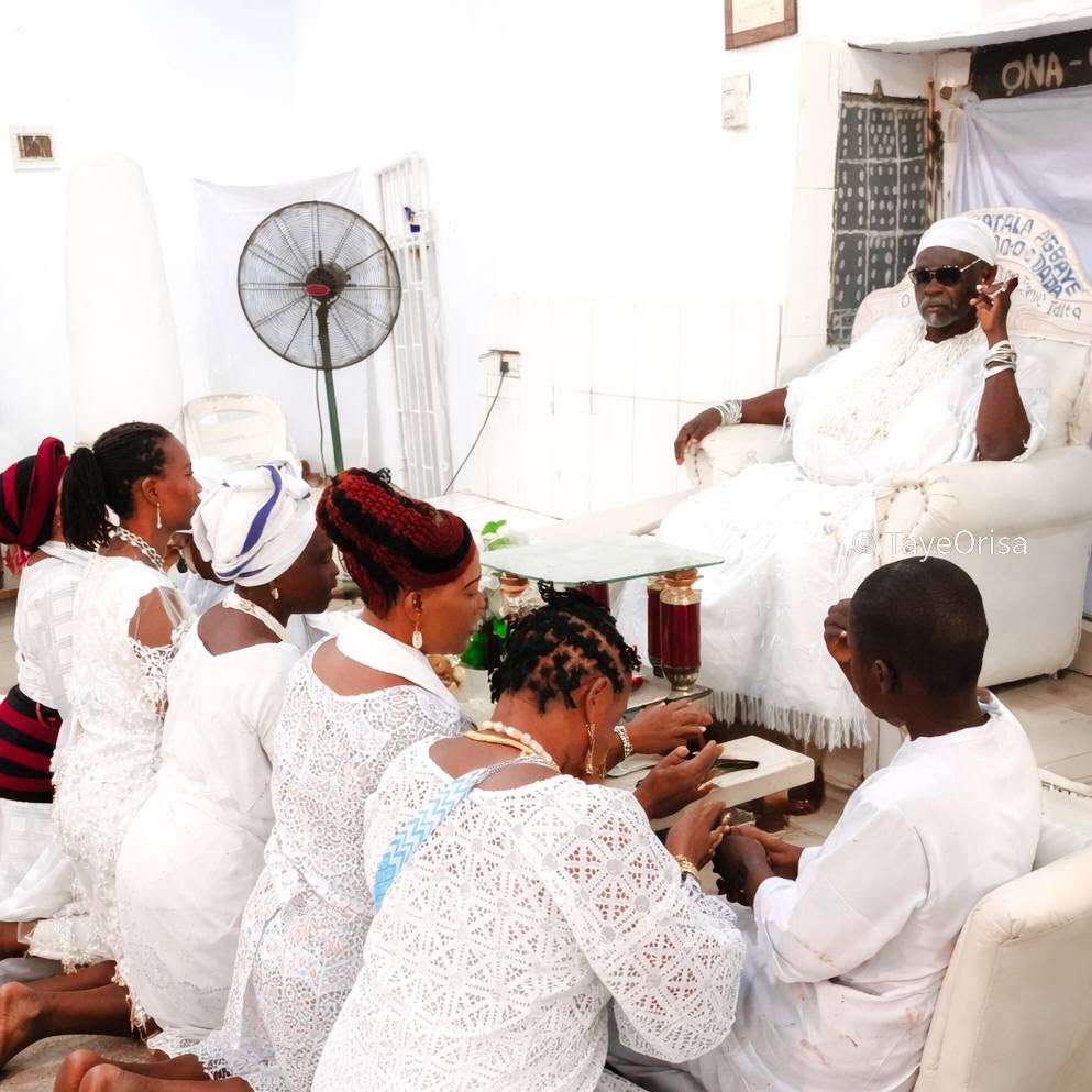 A seated elder in white Obàtálá regalia receiving visitors in the temple