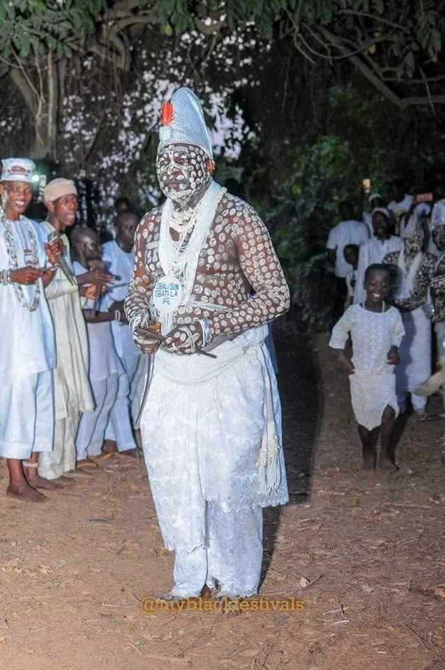 A ceremonial Obàtálá figure in white raiment, body markings and pointed cap, during a night festival procession
